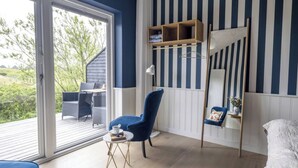 A room with a large sliding glass door leading to an outdoor deck, a blue chair, a small table with a cup, a wooden shelf with folded clothes, and a mirror on the wall. - Kellers Badehotel (Fano)