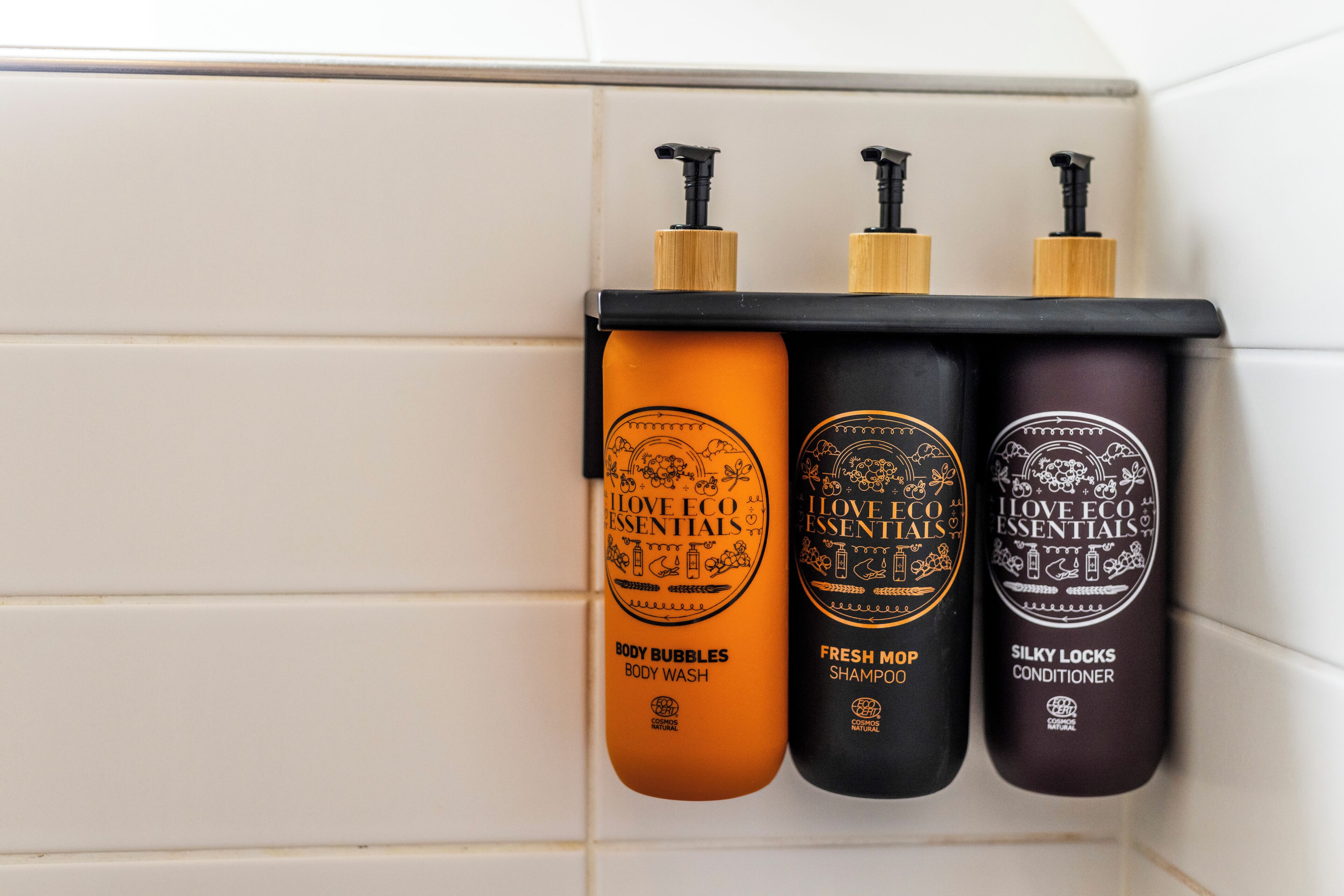 Tres botellas de I Love Eco Essentials sobre una pared de baldosas blancas.