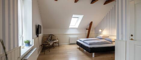 A bedroom with a sloped ceiling, a skylight, a bed, a chair, a table, and a television.