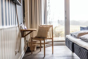 A room with a wooden chair, a small table, a bed with pillows, and a view through a large window. - Kellers Badehotel (Fano)