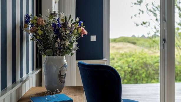 A room with a wooden desk, a vase with flowers, a blue chair, and a view of greenery outside. - Kellers Badehotel (Fano)