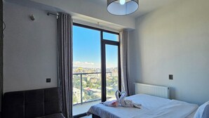 Un dormitorio moderno con una cama grande, un sofá y vista a un paisaje urbano a través de un ventanal.