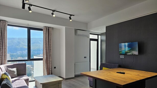 Una sala de estar moderna con un sofá, una mesa de centro y una mesa de comedor de madera. Hay un televisor montado en la pared y una ventana con cortinas.