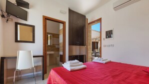 A hotel room with a red bedspread, a white chair, a desk, a TV, and a wardrobe. - Lido Marini Rooms Affittacamere B&B (Salve)