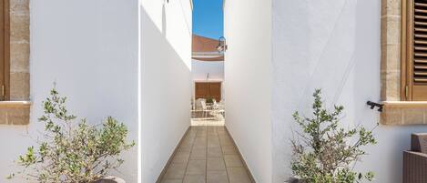 A narrow alleyway with potted plants on either side, leading to a patio area with outdoor furniture.