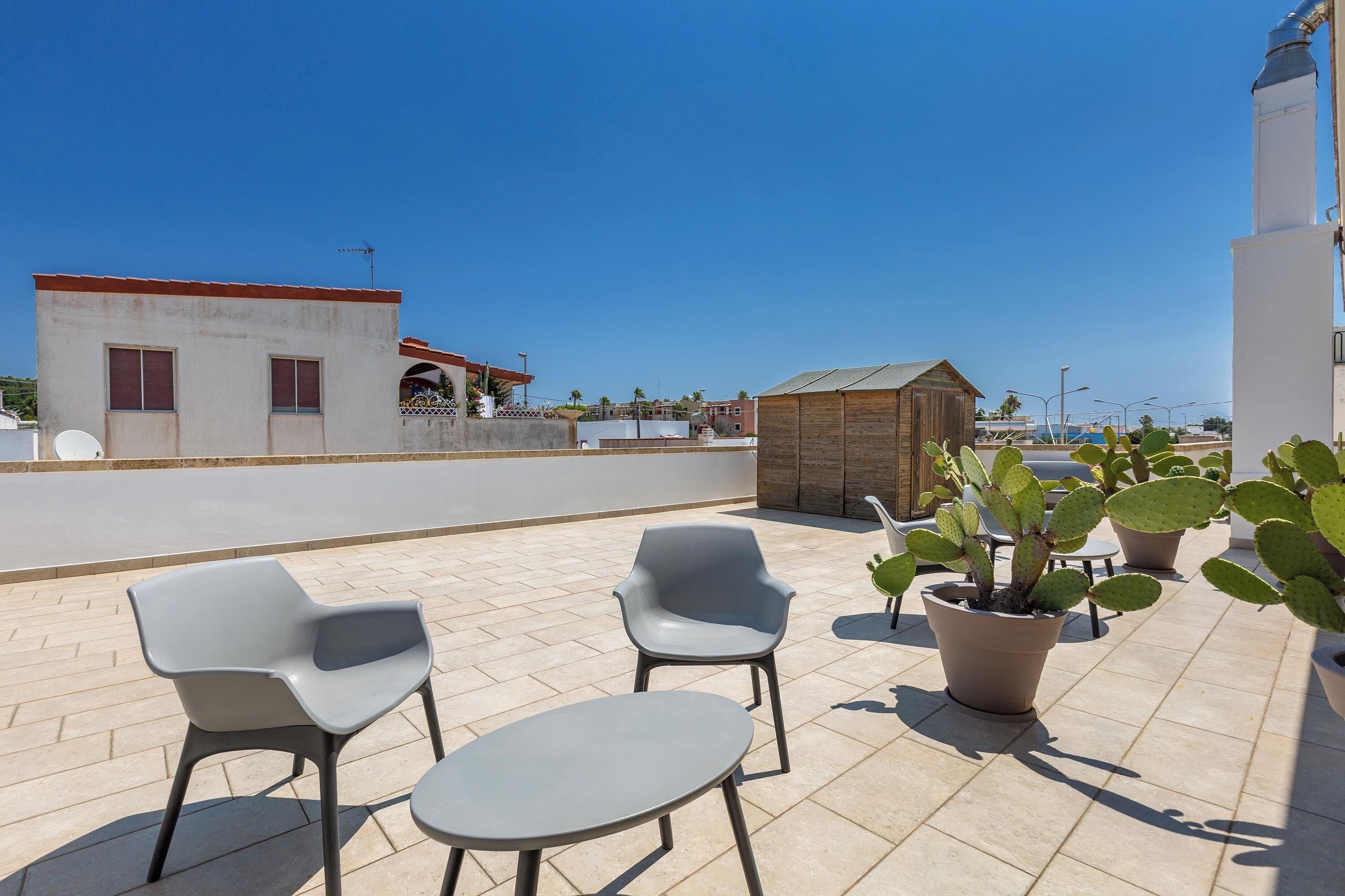 Une terrasse extérieure avec deux chaises grises et une petite table, entourée de cactus en pot, sous un ciel bleu dégagé.