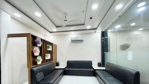 A modern waiting area with a black leather couch, a wooden shelving unit with decorative items, and an air conditioning unit on the wall. - Hotel SRK - near Temple  (Varanasi)