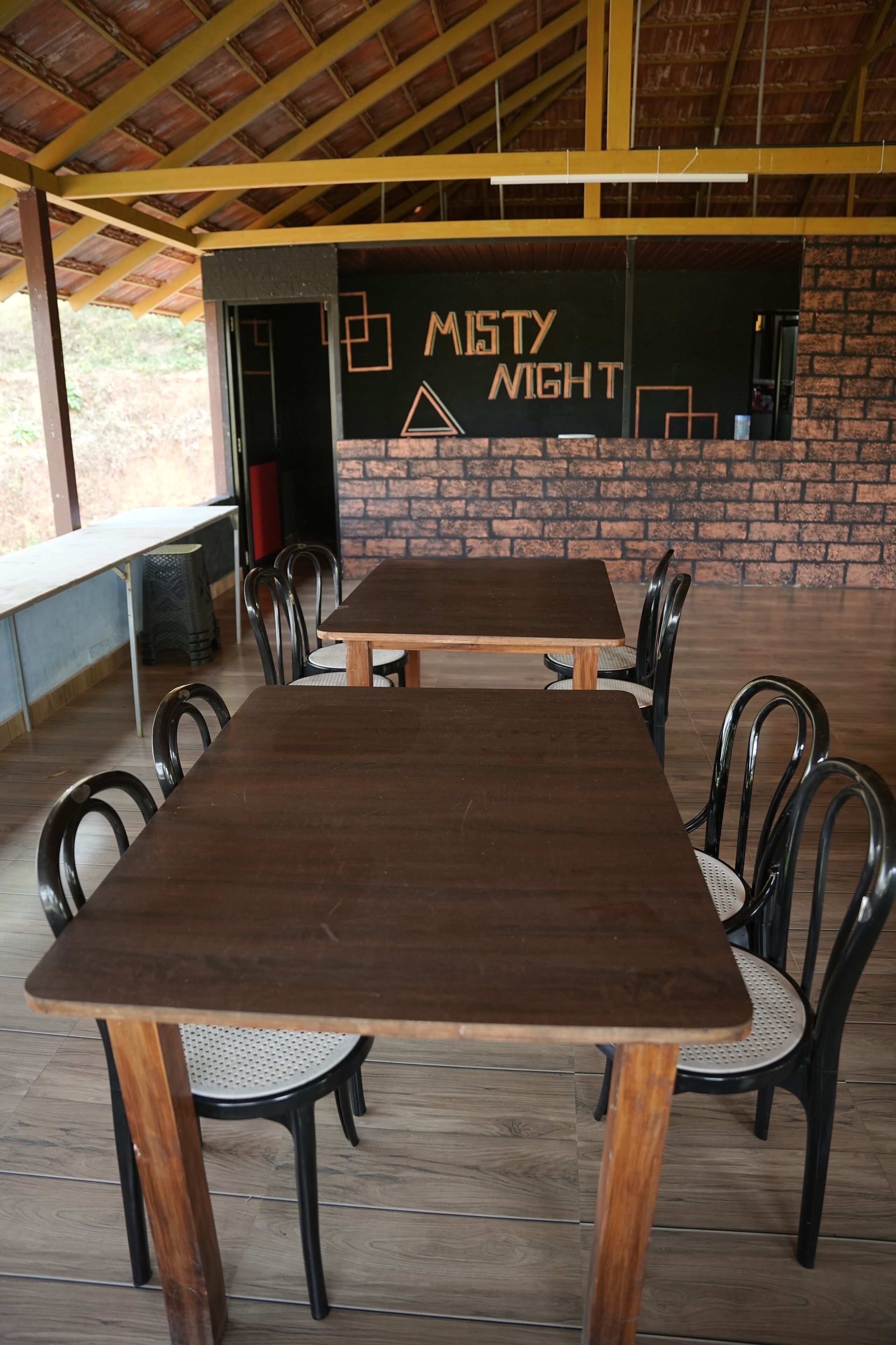 Een overdekt buitenterras met houten tafels en stoelen, een bakstenen muur en een bordje met de tekst 'MISTY NIGHT'.