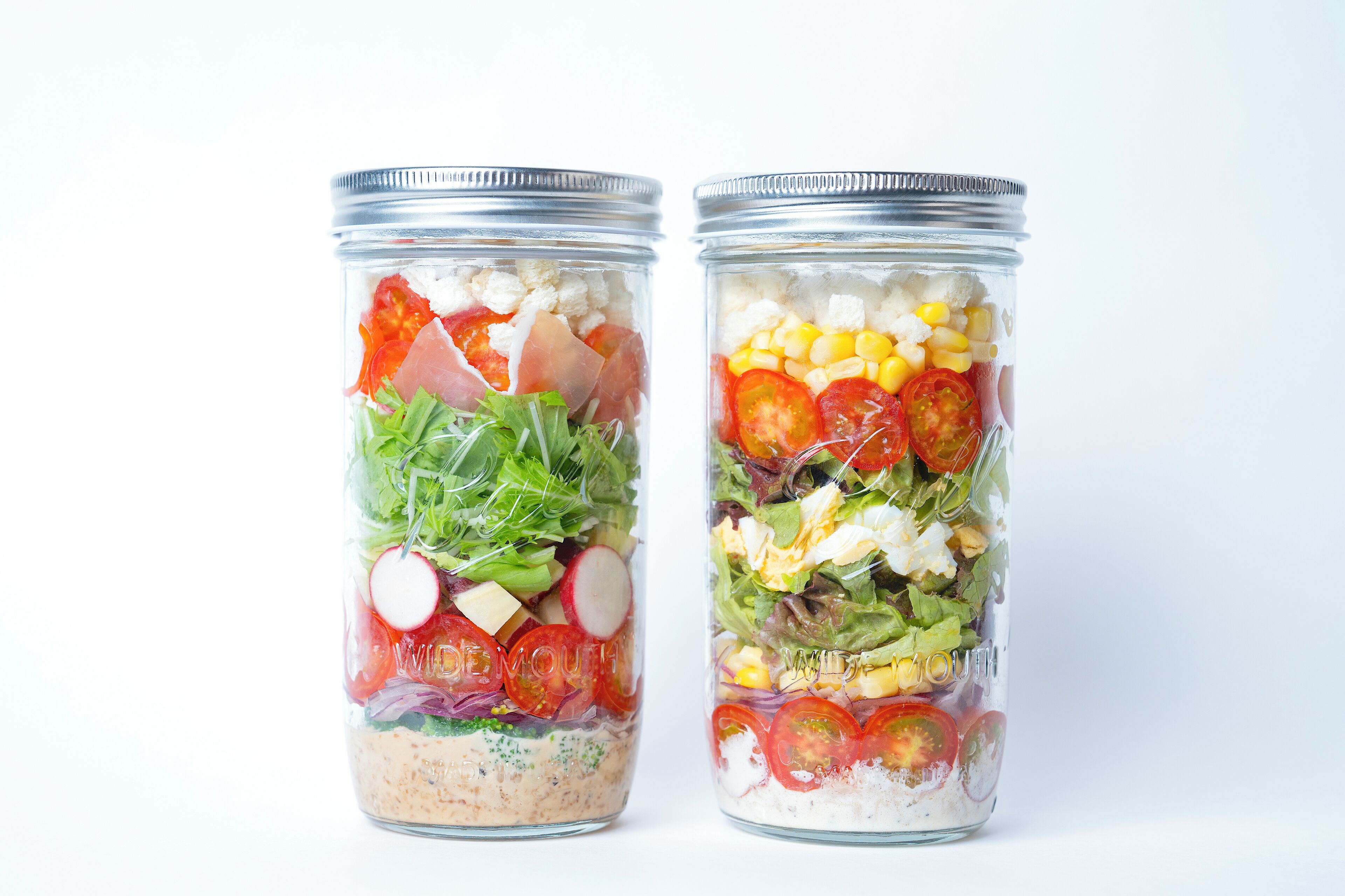 Dos frascos de vidrio con tapas metálicas, cada uno con una ensalada en capas y diversos vegetales y queso.