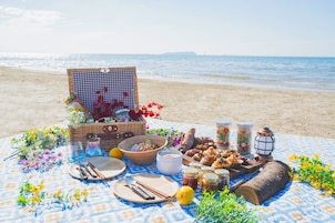 Un montaje para picnic en la playa con una manta a cuadros, comida, bebidas y un farol.