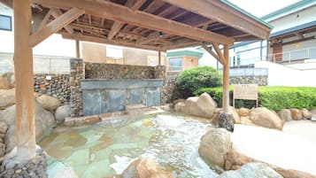 Un área de aguas termales de piedra con una pérgola de madera encima, rodeada de rocas y un muro de piedra.