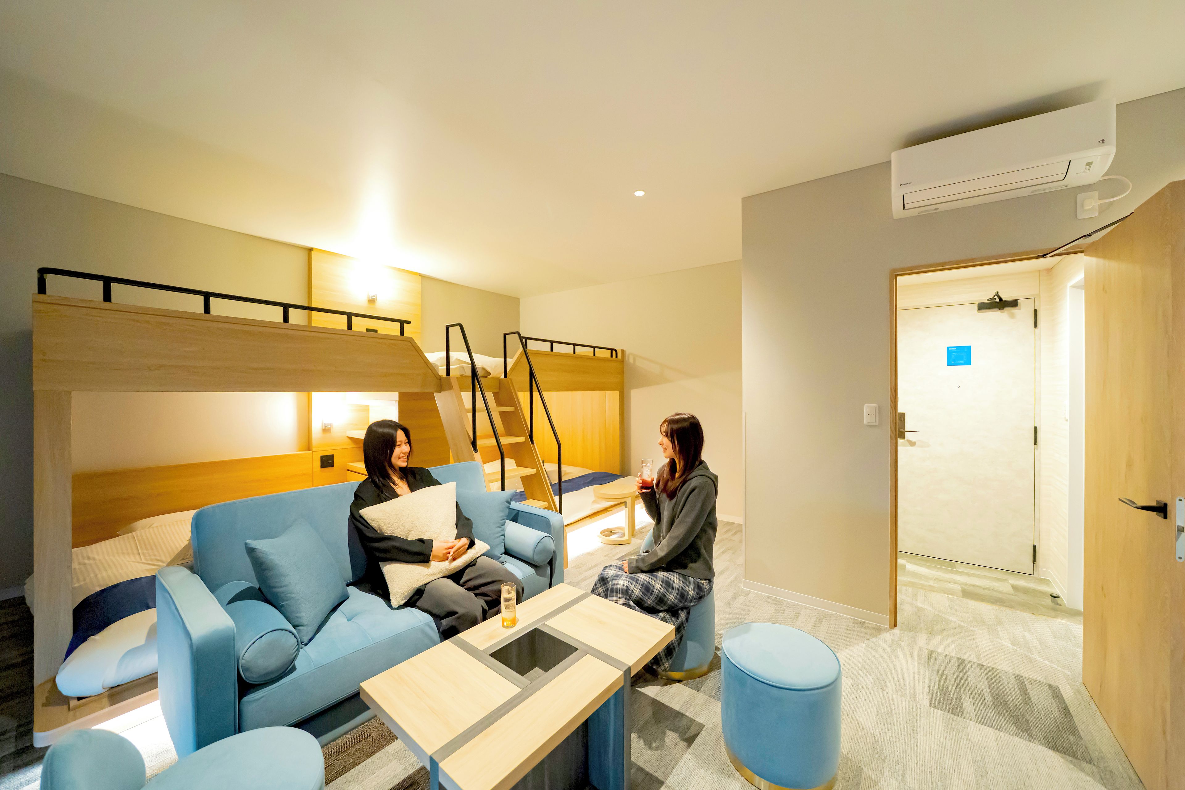 A modern hotel room with a sofa, a small table, a blue ottoman, and a staircase leading to an upper level.