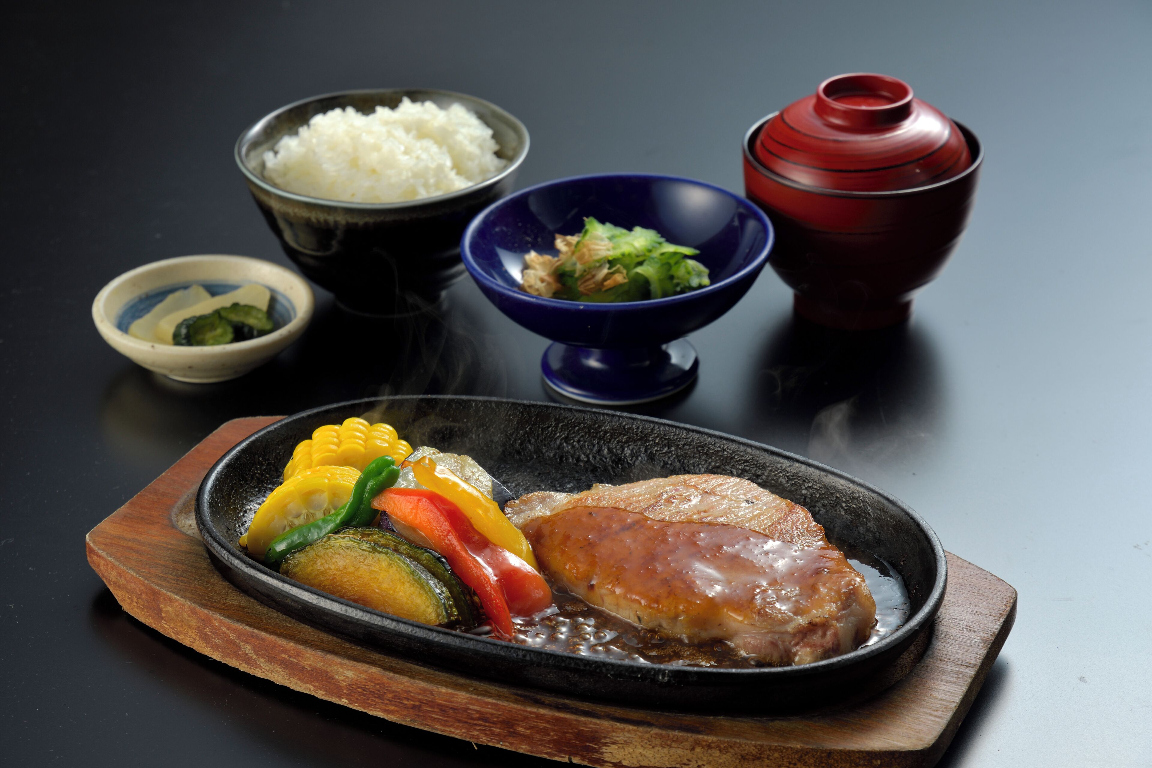 Un plato con arroz, verduras y carne sobre una mesita oscura, acompañado de cuencos y una olla roja.