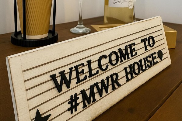 En träskylt med texten ”VÄLKOMMEN TILL #MWR HOUSE” står på ett bord.