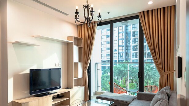 A modern living room with a flat-screen TV, a glass coffee table, a sofa, and a view of a high-rise building with greenery outside. - VinHomes Skylake Apartment My Dinh Nam Tu Liem (Hanoi)