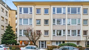 Een meerlaags woongebouw met een beige gevel, diverse ramen en een parkeerplaats met auto's.