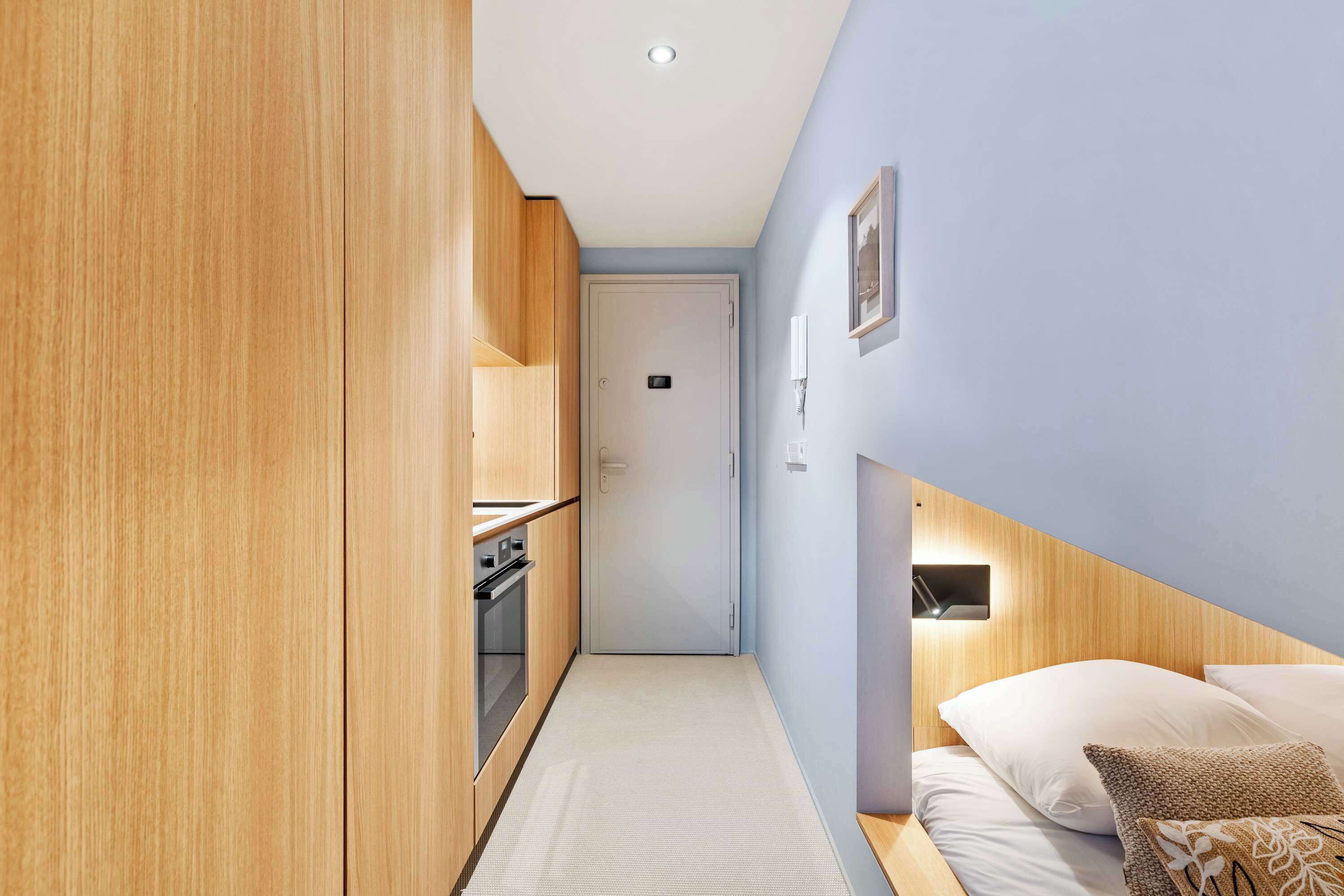 A narrow hallway with wooden cabinets, a white door, and a bed with white and patterned pillows.