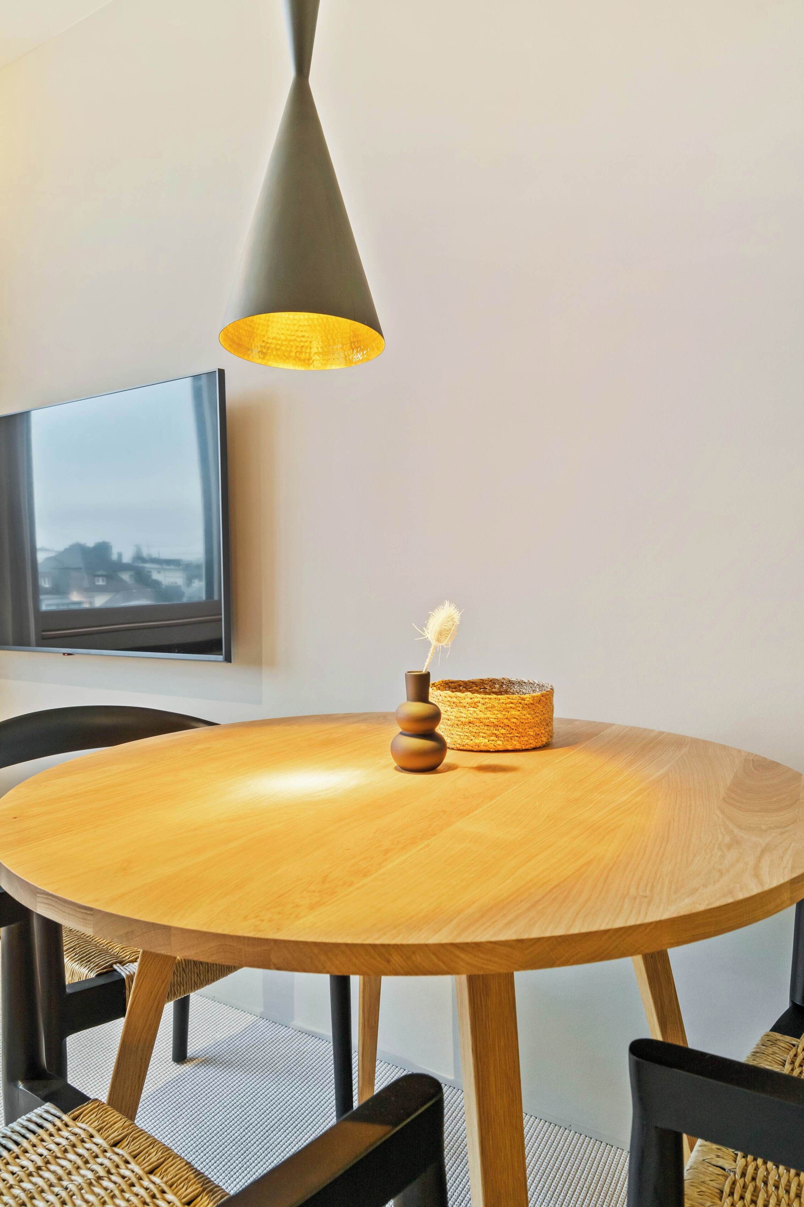 Une table ronde en bois, sur laquelle se trouvent un panier tressé et un vase contenant une fleur séchée.