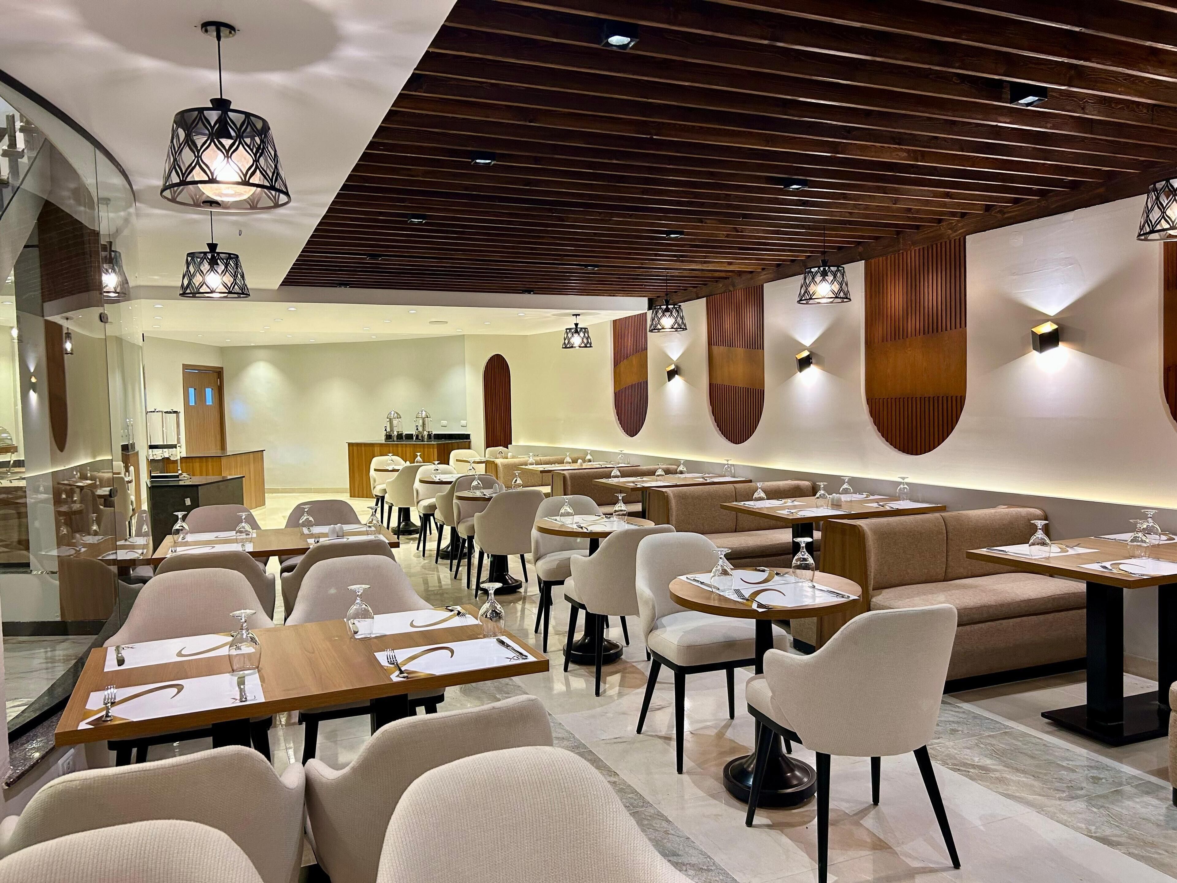 A modern restaurant interior with multiple dining tables and chairs, wooden ceiling beams, and pendant lighting.