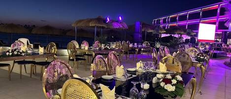 An outdoor dining area with tables set for a meal, surrounded by thatched umbrellas and beach views.