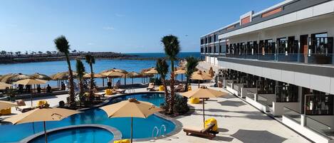 A pool area with sun loungers and umbrellas, a clear view of the sea, and a modern building with balconies.