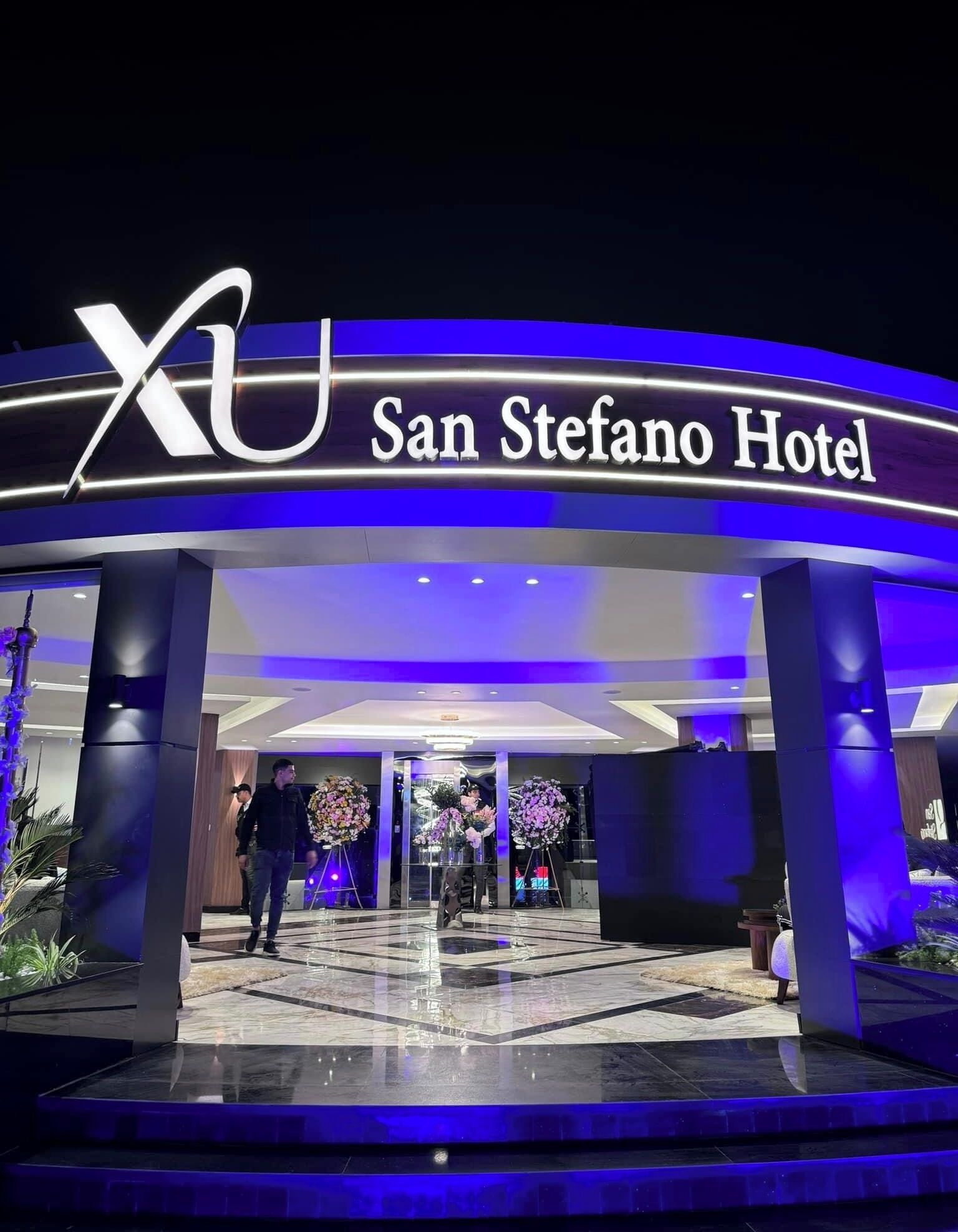 La entrada del Hotel San Stefano, con un llamativo logotipo 'XU' y un diseño moderno con iluminación azul.