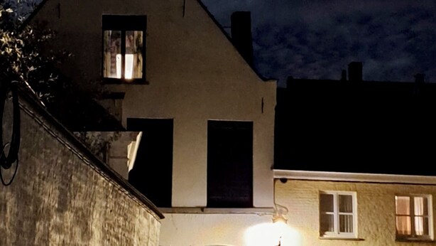 Une maison à deux étages, dotée d’un toit à forte pente, d’une cheminée bien visible et d’une fenêtre éclairée.