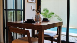 A dining area with a wooden table and chairs, a vase with flowers, and a view of a swimming pool through large windows. - Esencia Botanic Piscina Privada  (Tulum)