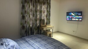 Een slaapkamer met een bed, een bureau, een televisie en een gordijn met een tropisch dessin.