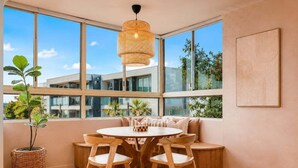A modern interior with a round wooden table, four chairs, a woven pendant light, and large windows offering a view of buildings and greenery.