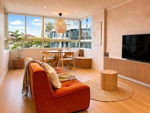 Ein modernes Wohnzimmer mit einer Couch, Esstisch, Stühlen, einem Fernseher und großen Fenstern mit Blick auf Gebäude und Bäume.