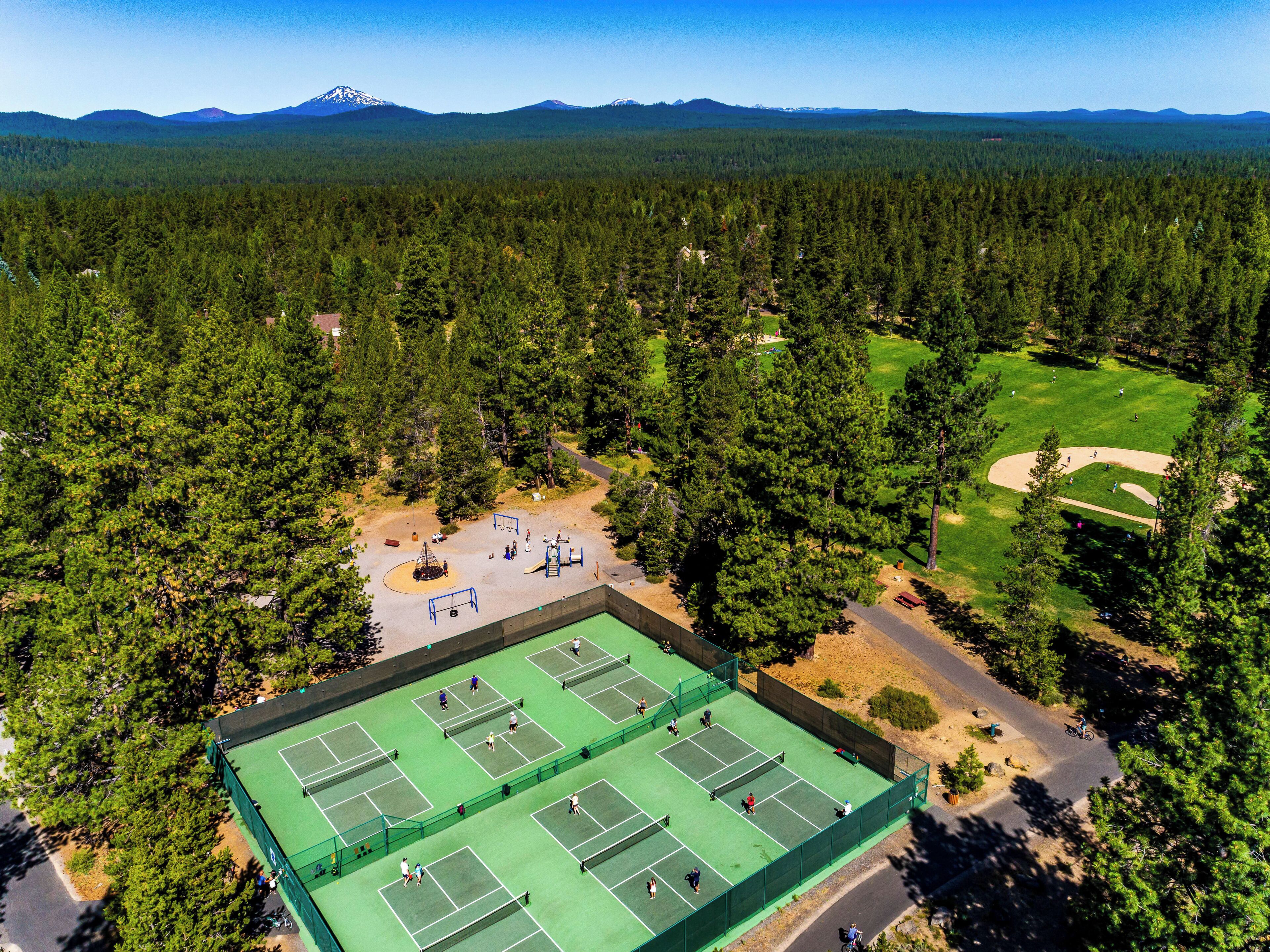 An aerial view of a tennis court surrounded by dense forest, with mountains in the background.