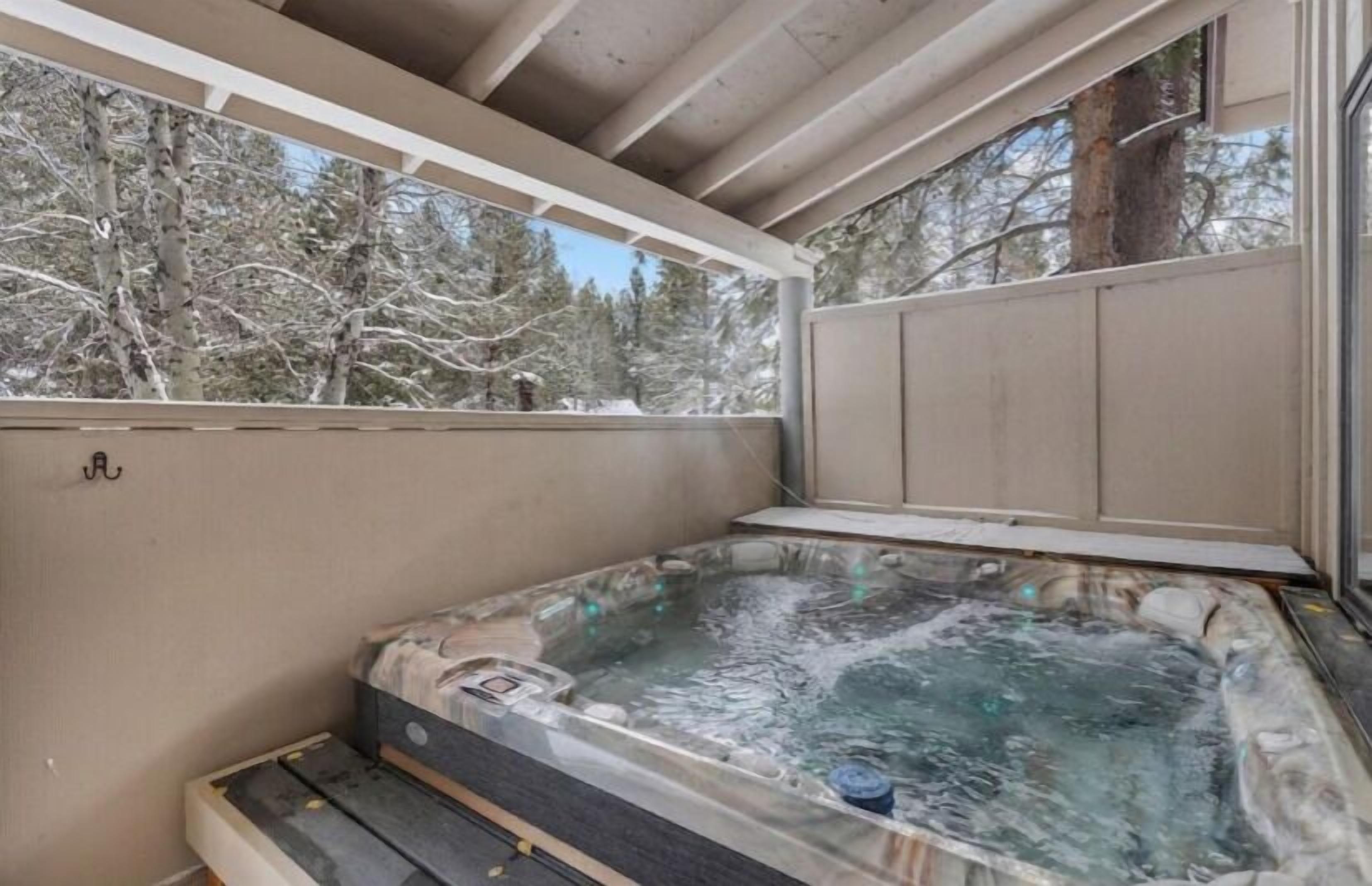 A hot tub with a wooden deck, surrounded by a wooden deck and a glass wall, with a view of snow-covered trees.
