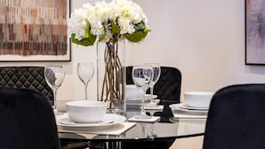 Uma mesa de jantar posta com pratos, tigelas e copos de vinho brancos, rodeada por cadeiras pretas.