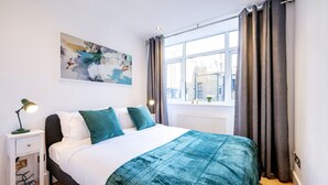 Quarto moderno com uma cama grande, mesa de cabeceira com luminária, janela com cortinas e vista para a cidade.