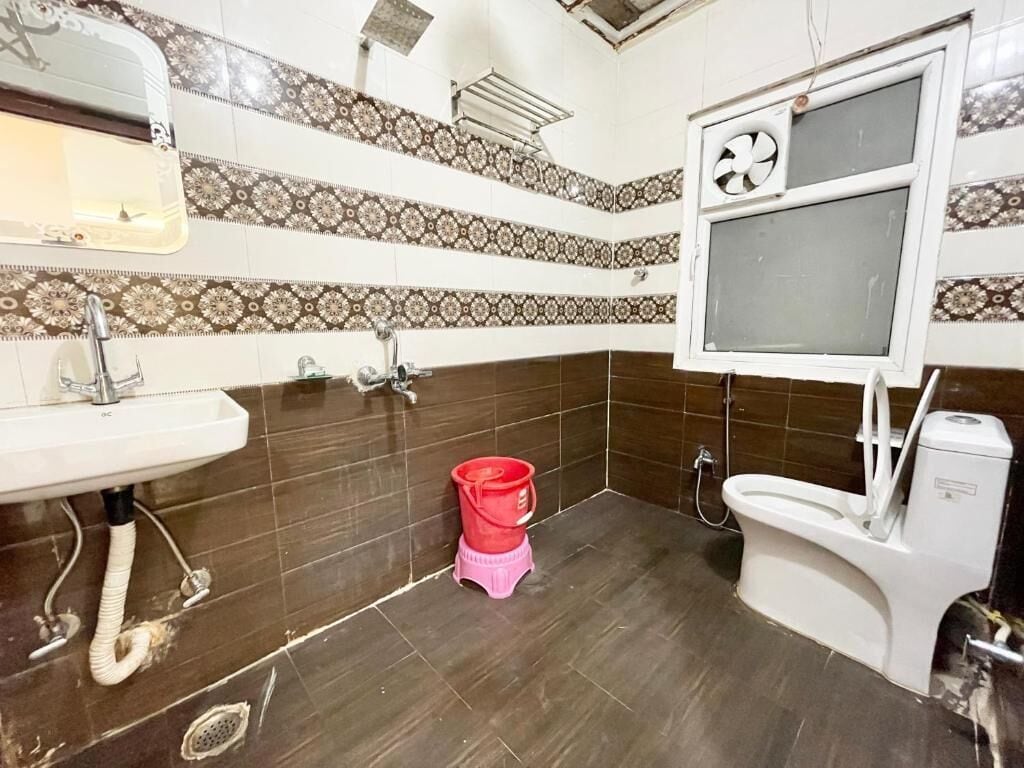 Une salle de bain avec un lavabo, des toilettes et une douche. Les murs sont recouverts de carreaux décoratifs, et il y a une fenêtre avec un ventilateur.