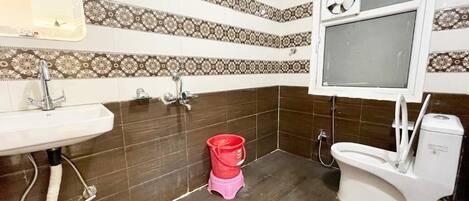 Une salle de bain avec un lavabo, des toilettes et une douche. Les murs sont recouverts de carreaux décoratifs, et il y a une fenêtre avec un ventilateur.