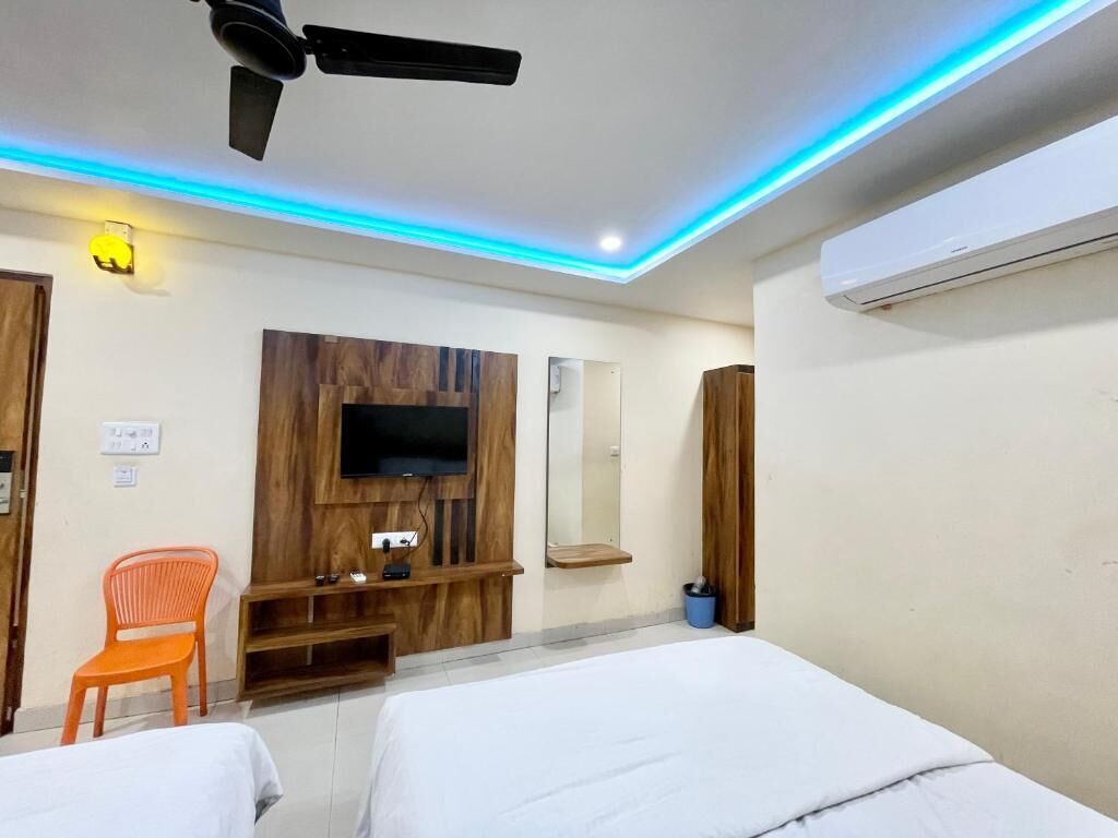 A hotel room with a ceiling fan, a flat-screen TV mounted on a wooden panel, a bed, a chair, and an air conditioning unit.