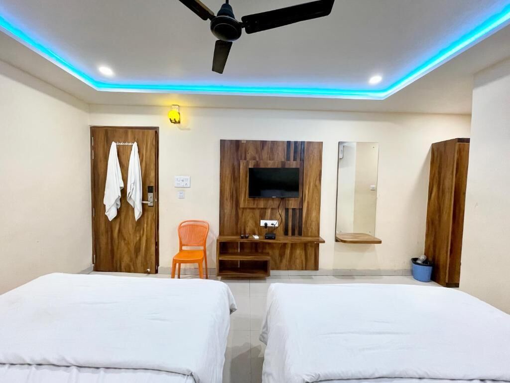 Une chambre d’hôtel avec deux lits, un ventilateur de plafond, un téléviseur fixé sur un panneau en bois, un fauteuil orange et une armoire en bois.