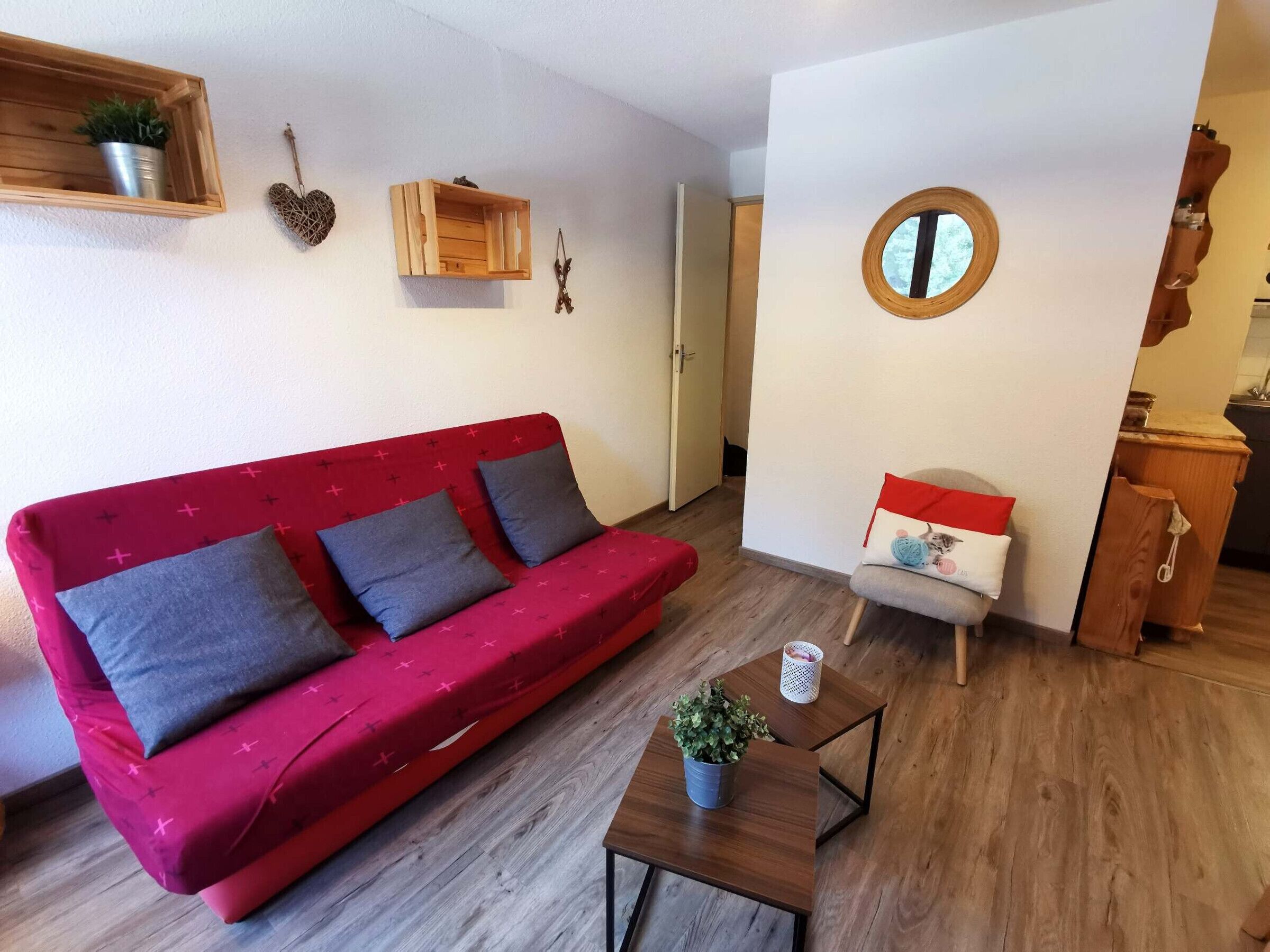 En röd soffa med gråa kuddar, ett litet träbord med en växt och en rund spegel på väggen.