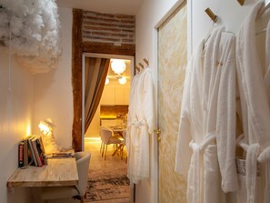 A room with a wooden desk, books, a chair, a door with towels, and a glimpse of a kitchen in the background.