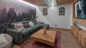 A modern living room with a grey sofa, colorful cushions, a wooden coffee table, and a forest mural on the wall.