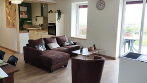 A modern living room with a brown leather sofa, a small table, and a chair. There is a kitchen in the background with white cabinets and a green backsplash. A clock is on the wall, and there are windows with curtains. - Natural Cottage, Near Montluçon & Néris-les-Bains (Beaune-d'Allier)