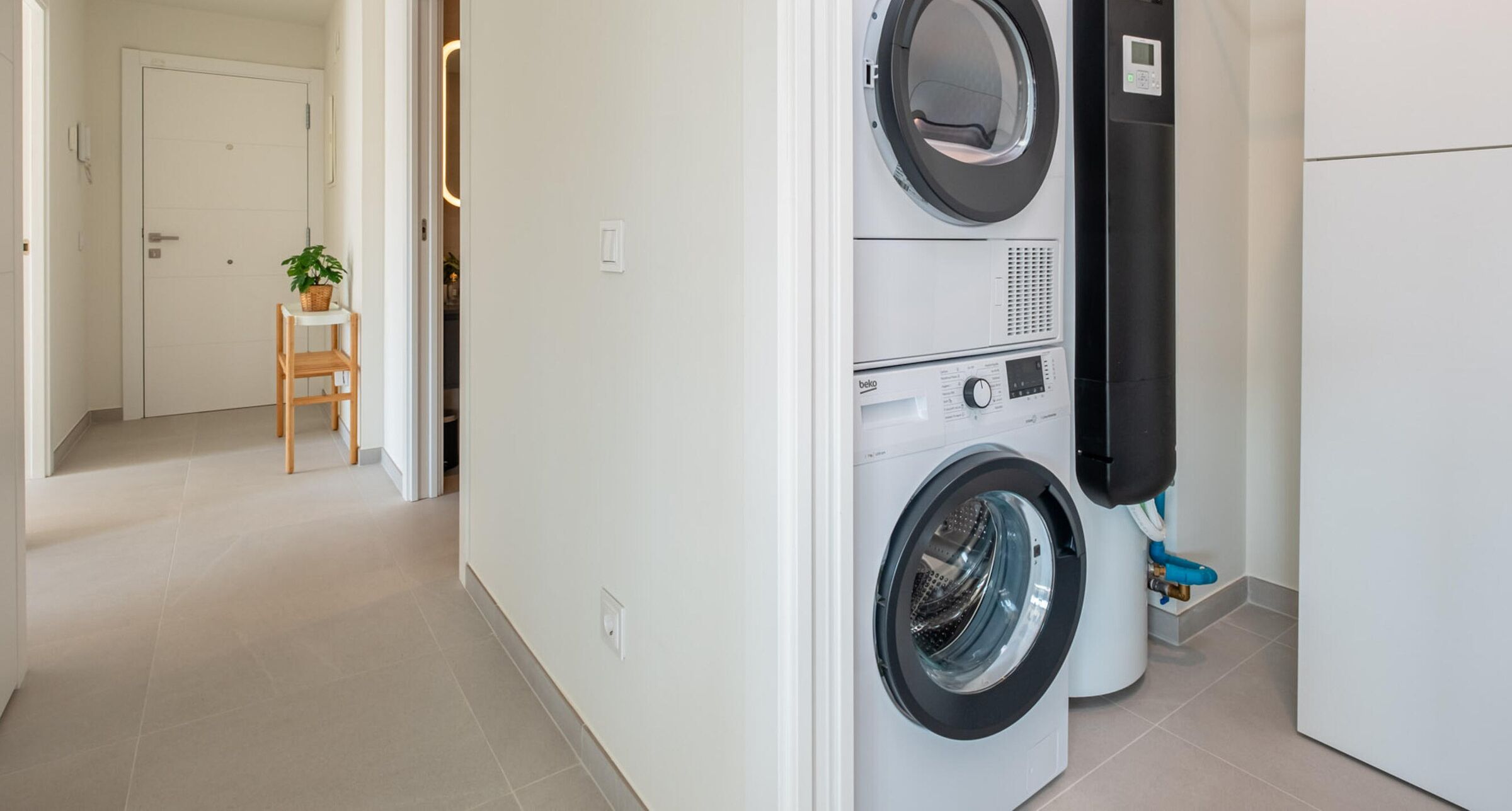 A laundry area with stacked washing machines, a small wooden stool, and a potted plant.