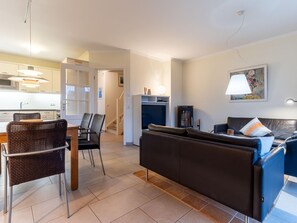 Un salon moderne avec un canapé, des fauteuils, un coin repas avec une table, et une cuisine en arrière-plan.
