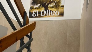 Egy bézs színű lépcsőfokokkal és fa korláttal ellátott lépcső vezet egy falhoz, amelyen egy kép van, a képen egy fa látható, és a kép mellett a „El Olivo” felirat szerepel.