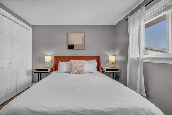 Une chambre d’hôtel avec un grand lit, deux tables de chevet avec des lampes, une tête de lit, un tableau encadré au mur et une fenêtre avec des rideaux.