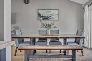 Een eetruimte met een houten tafel, grijs gestoffeerde stoelen, een bank, een vaas met takken en een schilderij aan de muur.