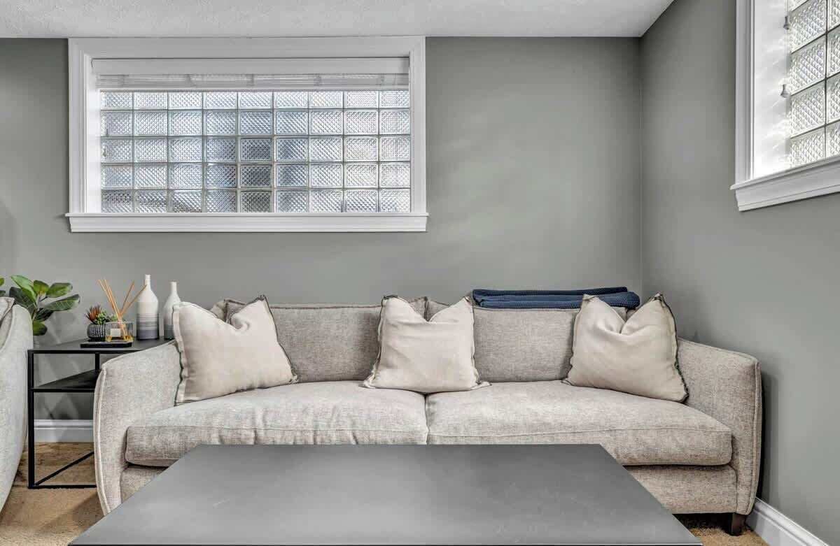 A living room with a grey sofa, a coffee table, a window with a grid pattern, and a small side table with decorative items.