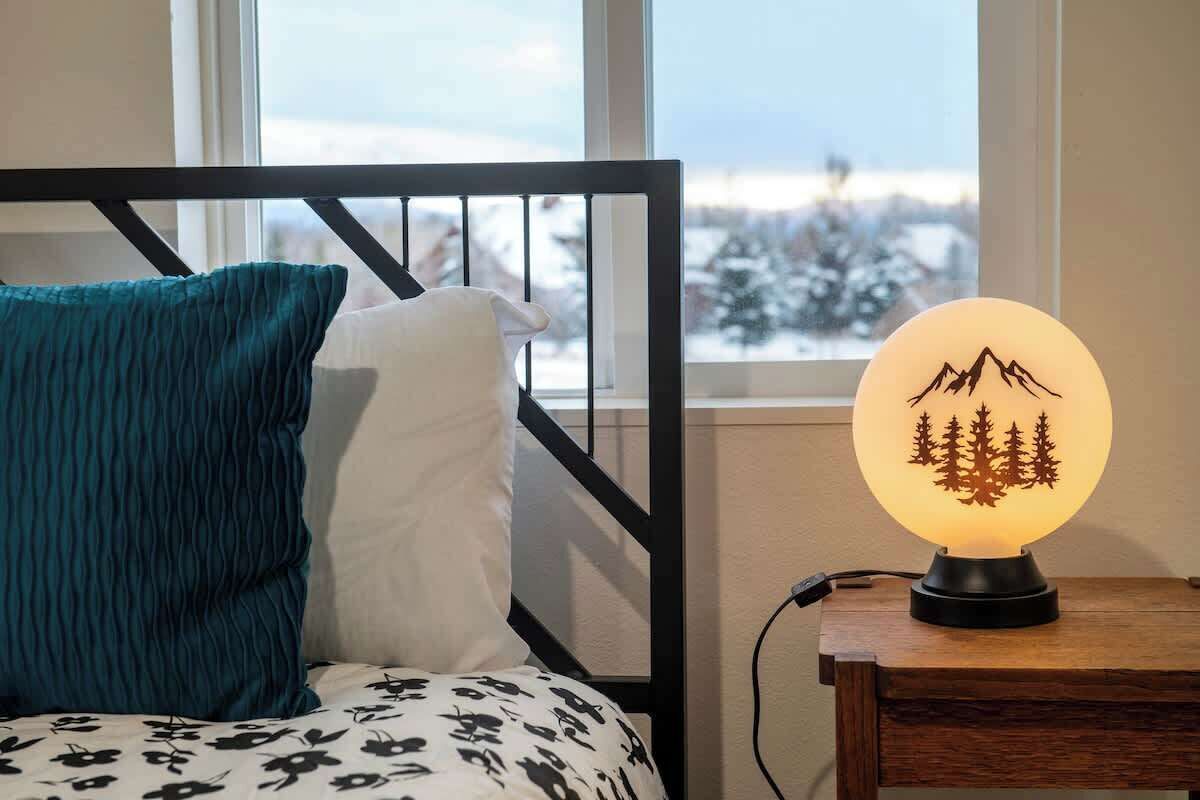 Ein Bett mit einem gemusterten Überwurf, einem türkisfarbenen Kissen und einem weißen Kissen. Eine Nachttischlampe mit Berg- und Baumkonturen steht auf einem hölzernen Nachttisch neben dem Bett.